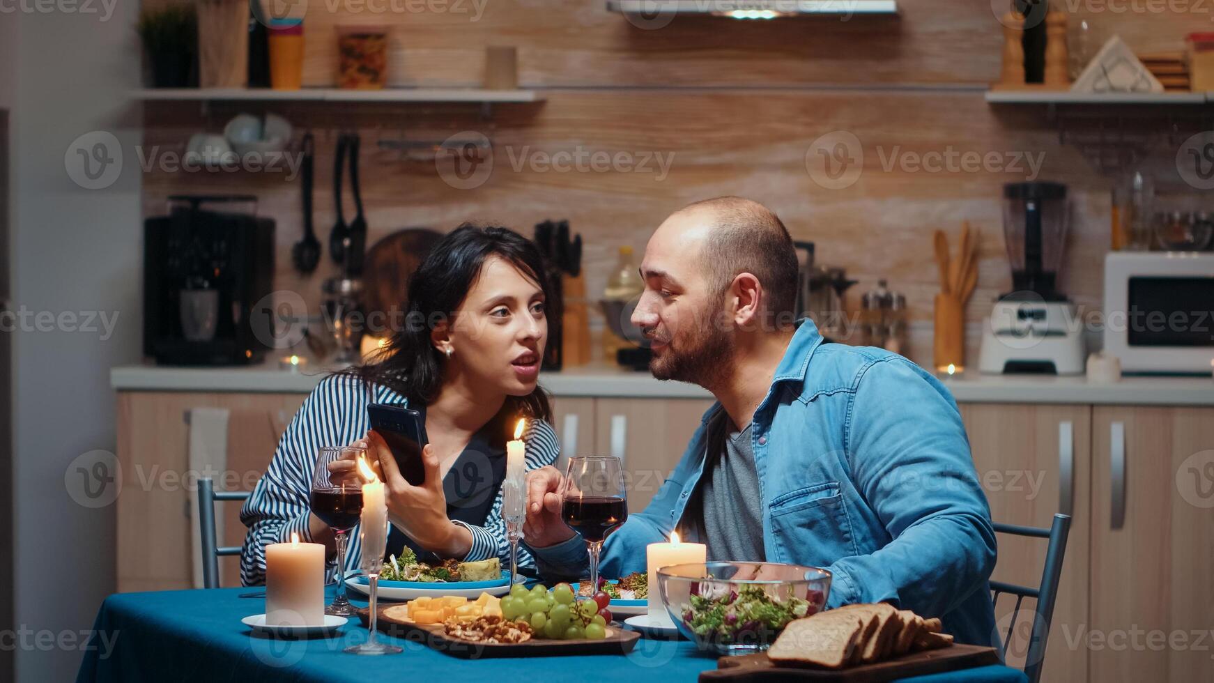 Young married couple using phone during romantic dinner. Adults sitting at the table in the kitchen browsing, searching, using smartphones, internet, celebrating their anniversary in the dining room. photo