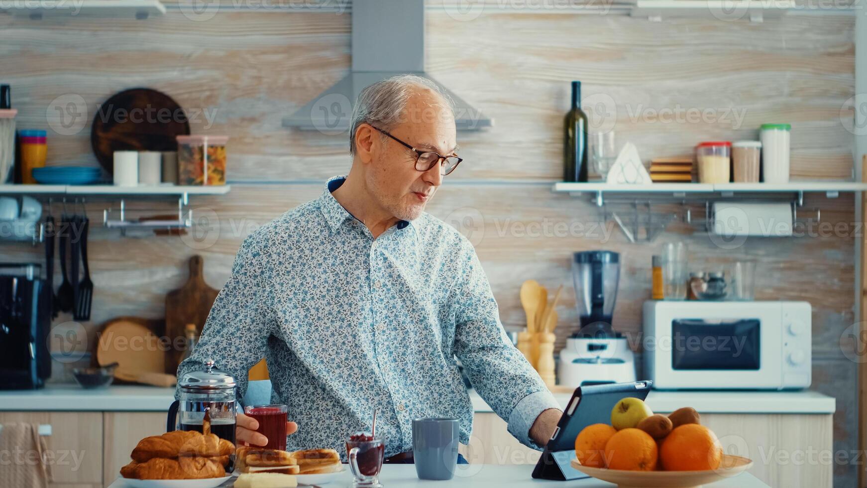Senior man using tablet computer while drinking tea during breakfast in kitchen. Elderly person with tablet portable pad PC in retirement age using mobile apps, modern internet online information technology with touchscreen photo