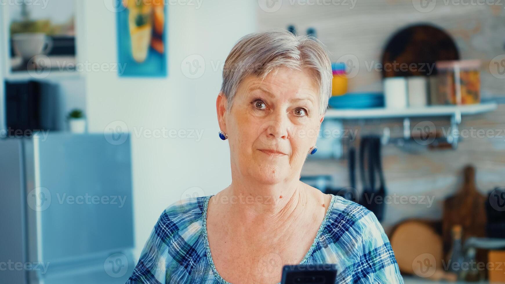 Granny looking at camera while using smartphone in kitchen during breakfast. Authentic elderly person using modern smartphone internet technology. Online communication connected to the world, senior leisure time with gadget at retirement age photo