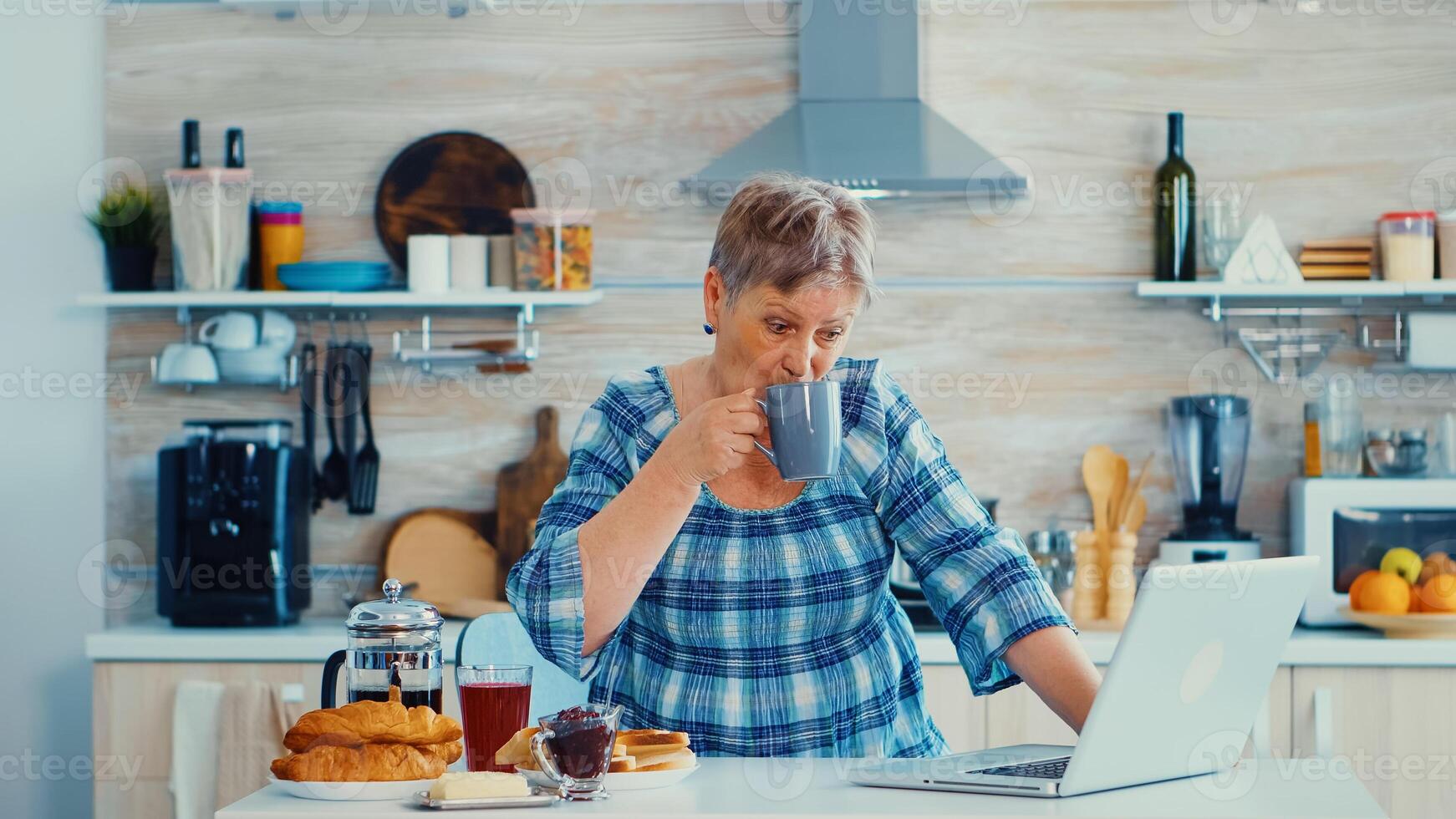 Senior businesswoman drinking coffee and working on laptop in kitchen during breakfast. Elderly retired person working from home, telecommuting using remote internet job online communication on modern technology notebook photo