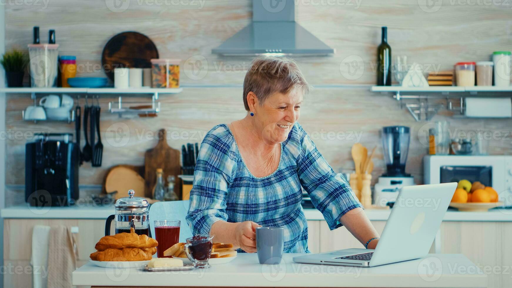 Senior woman drinking coffee and waving during a video conference with family in kitchen while having breakfast. Elderly person using internet online chat technology video webcam making a video call connection camera communication conference call photo