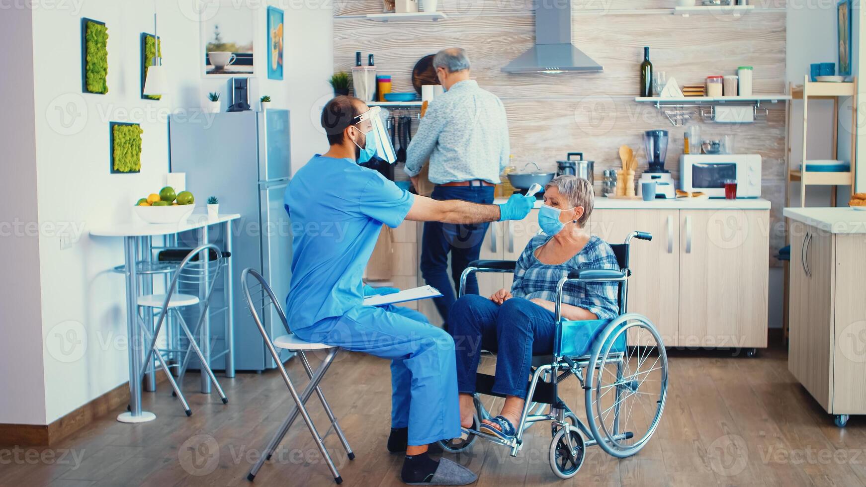 Temperature measurement using infrared thermometer on disabled senior woman in wheelchair during home visit in coronavirus pandemic. Social worker with visor and mask helping covid19 prevention photo