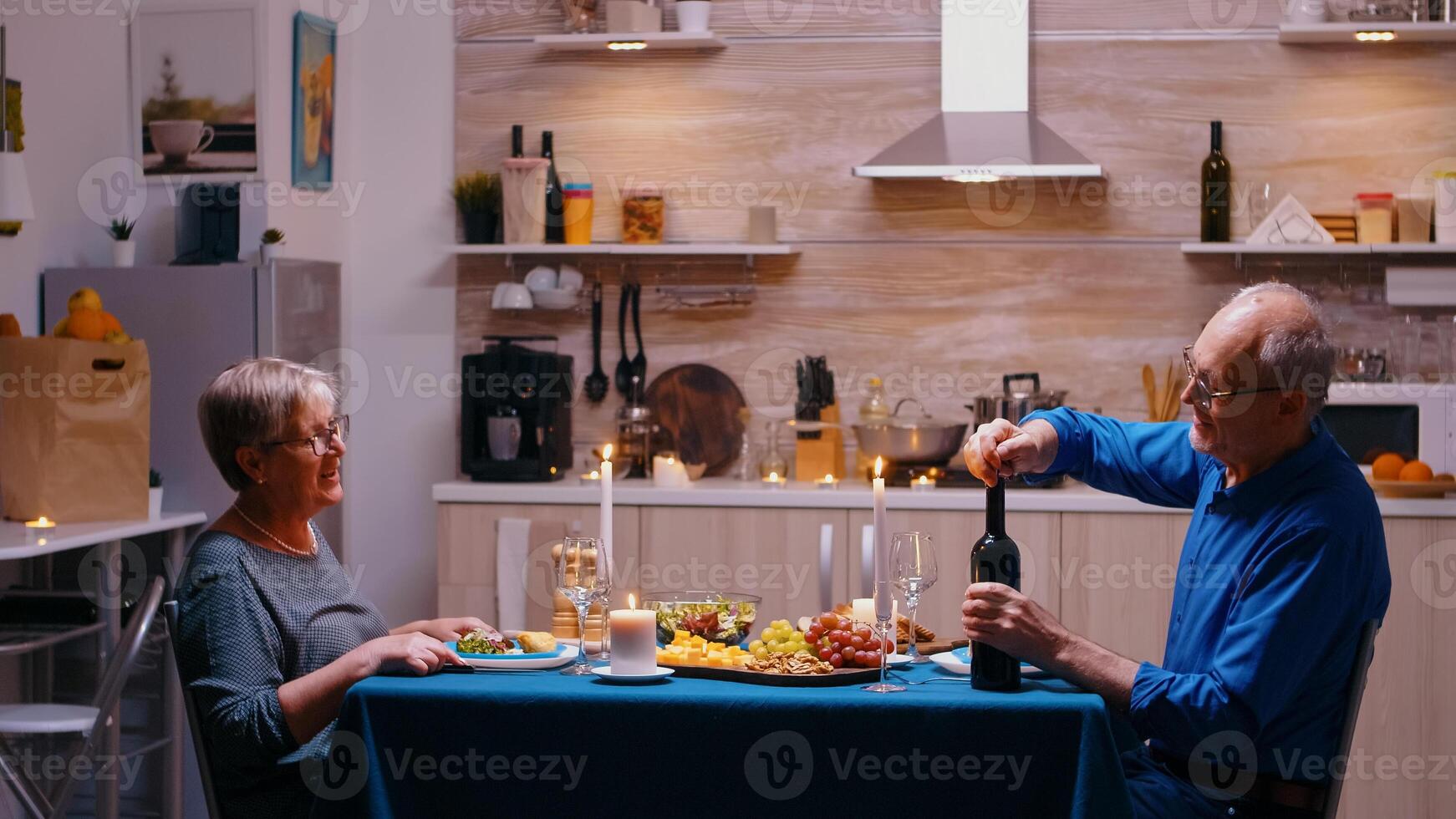 Elderly man opening a bottle of red wine during romantic dinner. Senior old couple talking, sitting at the table in kitchen, enjoying the meal, celebrating their anniversary at home with healty food. photo