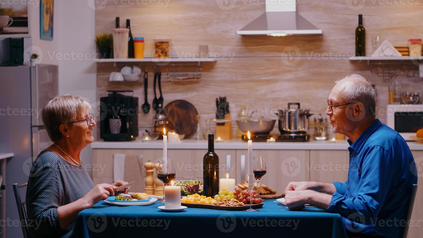 mayor Pareja relajarse comiendo cena y Bebiendo vino lentes juntos en el cocina a hogar teniendo un romántico cena. Jubilación Pareja celebrando su aniversario en el comida habitación. foto