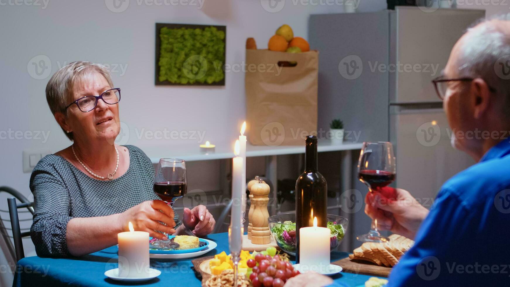 mayor relajado Pareja comiendo cena y Bebiendo lentes de rojo vino juntos en el cocina a hogar. anciano, retirado antiguo personas disfrutando el comida, celebrando su aniversario en el comida habitación. foto