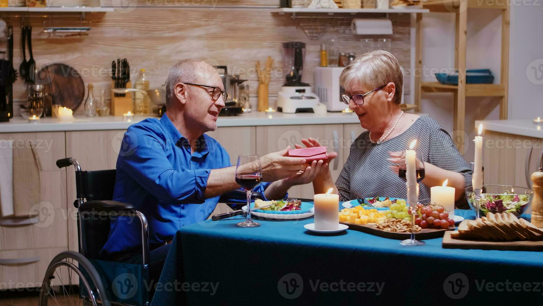 Senior cheerful woman holding a gift from her man sitting at the table in the kitchen during romantic meal. Imobilized paralyzed handicapped elderly husband dining with wife at home, enjoying the meal, celebrating their anniversary photo