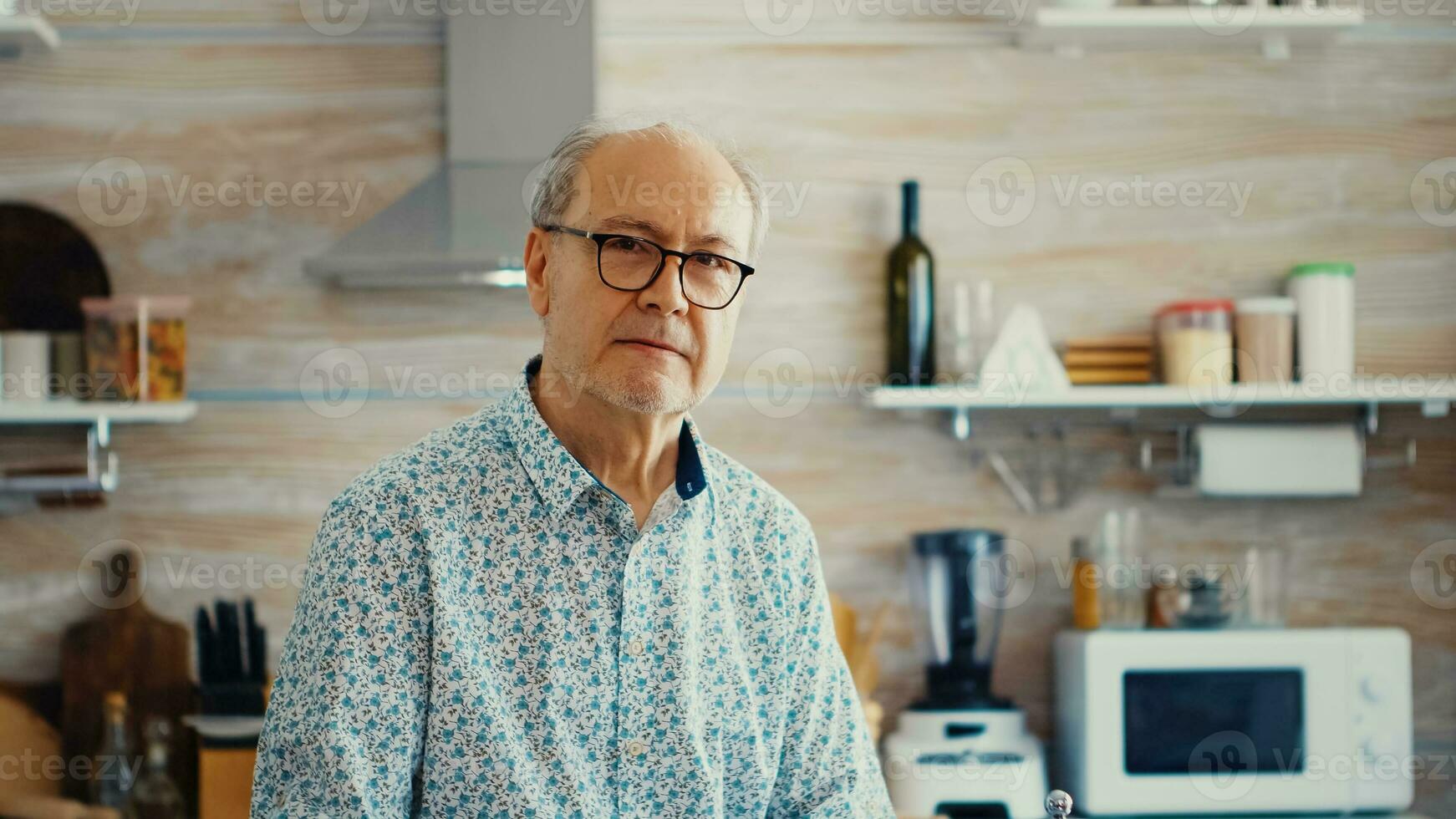 Mature man with wrinkles looking at camera in kitchen while enjoying a cup of coffee. Portrait of relaxed elderly senior older person in the morning, enjoying fresh warm drink. Healthy smiling adult face photo