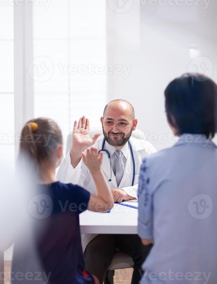 Physician doctor smiling at child and giving highfive during consultation in hospital office. Pediatrician specialist in medicine providing healthcare services treatment examination to family photo