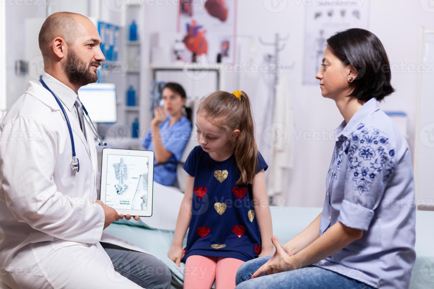 Pediatrician doctor explaining bones disease to mother using tablet with schelet image for sick child in hospital office. Physician specialist in pediatry medicine providing professional services photo