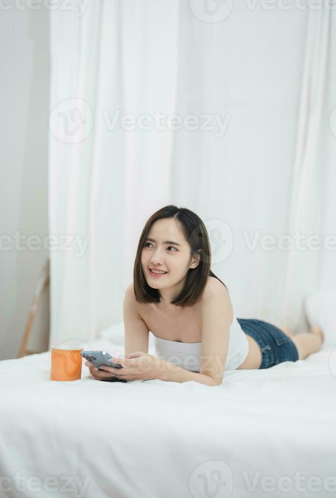 Cute Asian woman lying on bed using phone, exploring social media and sipping coffee in the morning. photo