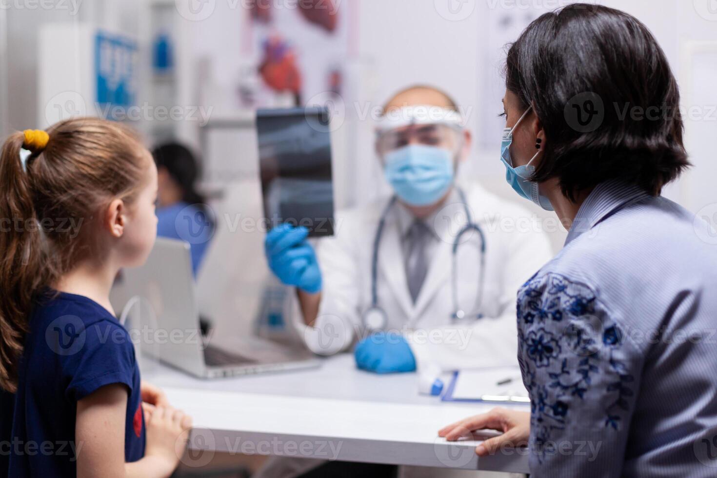 Caucasian doctor examining x-ray bones image of child and explaing treatment to prevent illness. Pediatrician specialist with protection mask and visor against covid19 providing healthcare service photo
