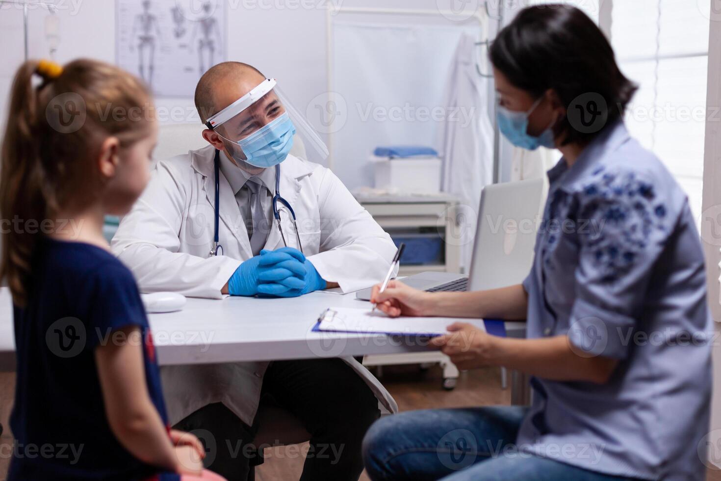Mother completing medical questionnaire at doctor office in hospital clinic. Pediatrician with protection mask and visor against covid19 talking about diagnostic resultes providing care services photo