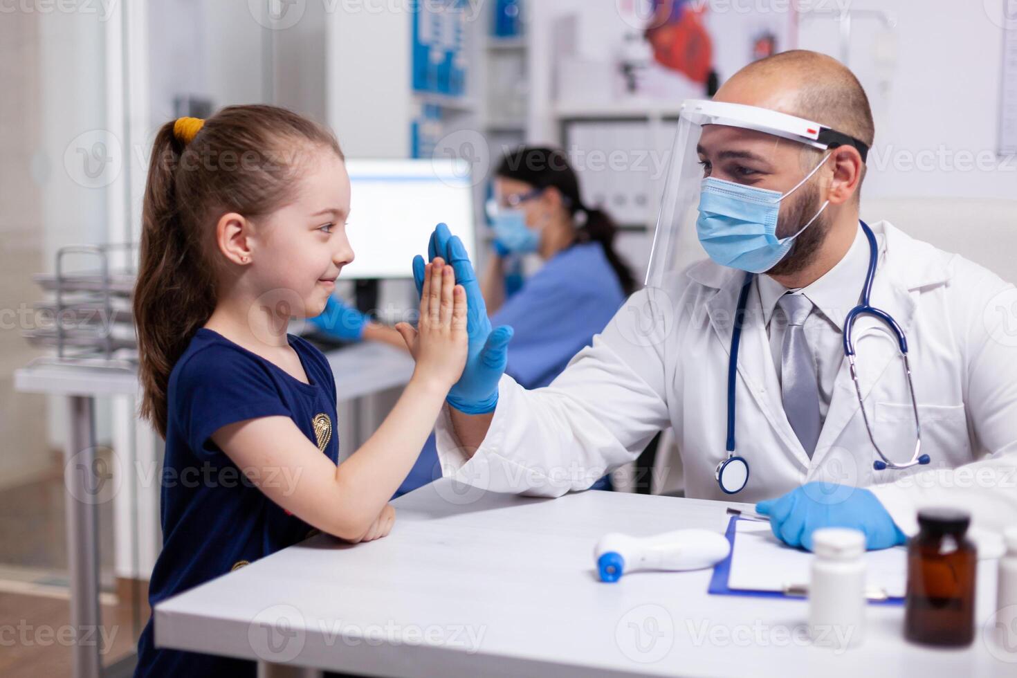 Pediatrician wearing face mask giving high-five and having conversation with child during expertise consultation. Pediatry doctor in protection gloves and visor providing care services in medical clinic photo