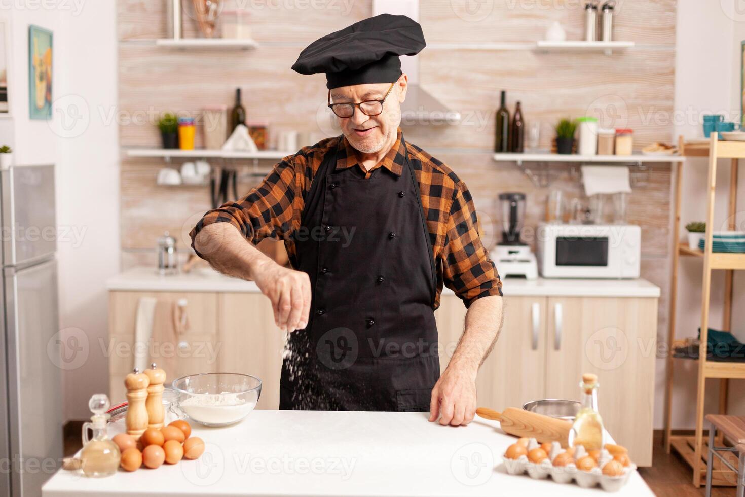 Senior chef preparing homemade bread sprinkling wheat flour on kitchen table. Retired senior chef with bonete and apron, in kitchen uniform sprinkling sieving sifting ingredients by hand. photo