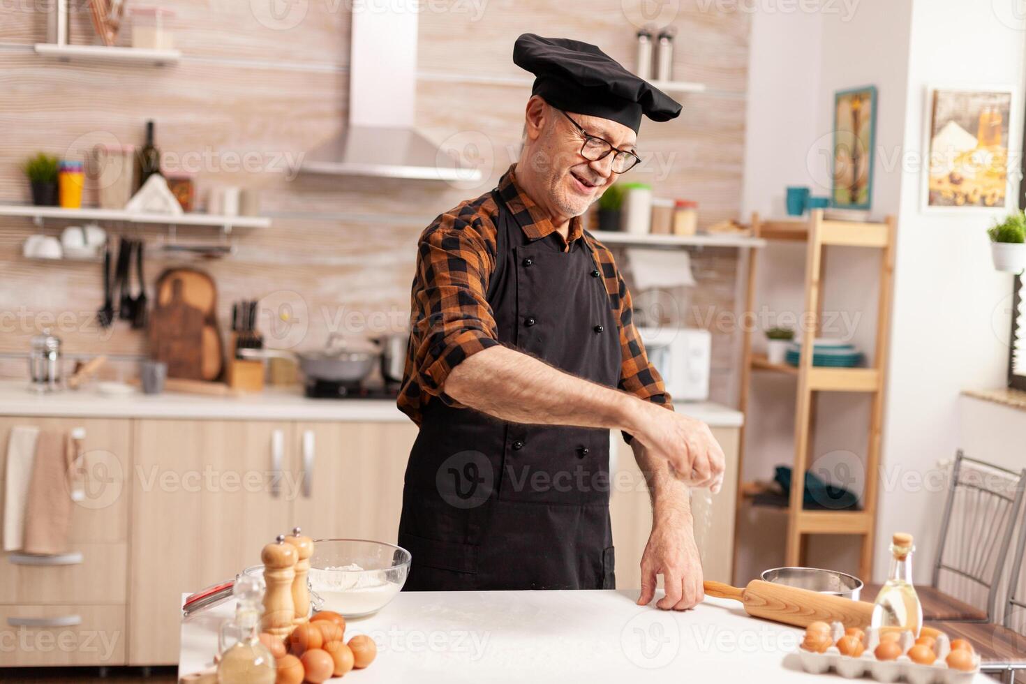 retirado cocinero en hogar cocina extensión trigo harina terminado mesa mientras preparando hecho a mano cocinar F con bonete y delantal, en cocina uniforme aspersión tamizado cernido ingredientes por mano. foto