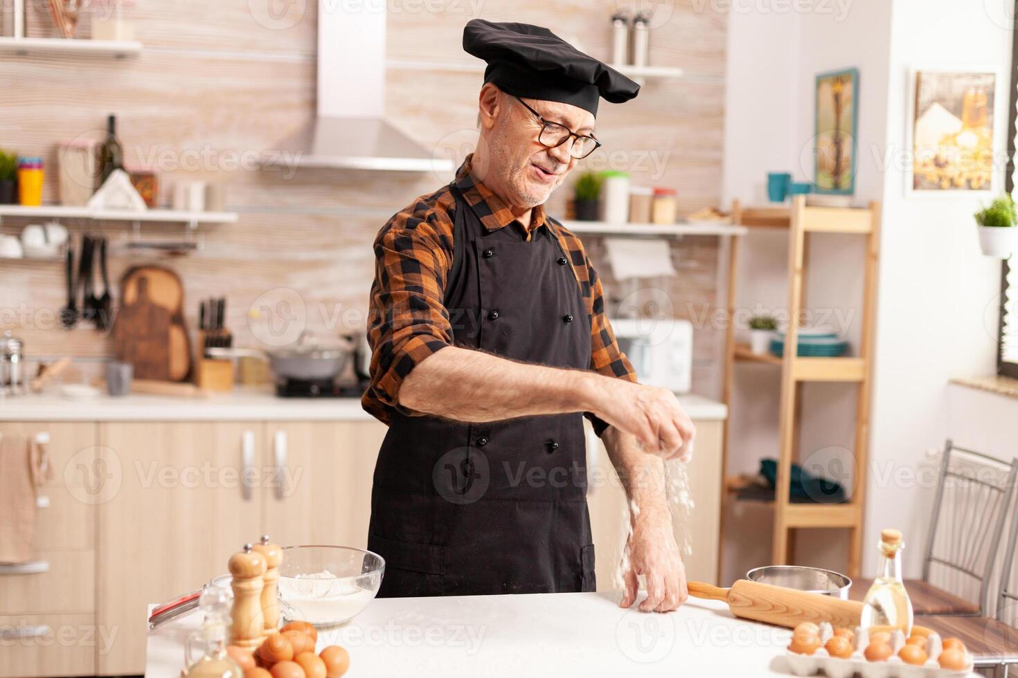 Experienced senior chef cooking healthy balanced food in home kitchen. Retired senior chef with bonete and apron, in kitchen uniform sprinkling sieving sifting ingredients by hand. photo
