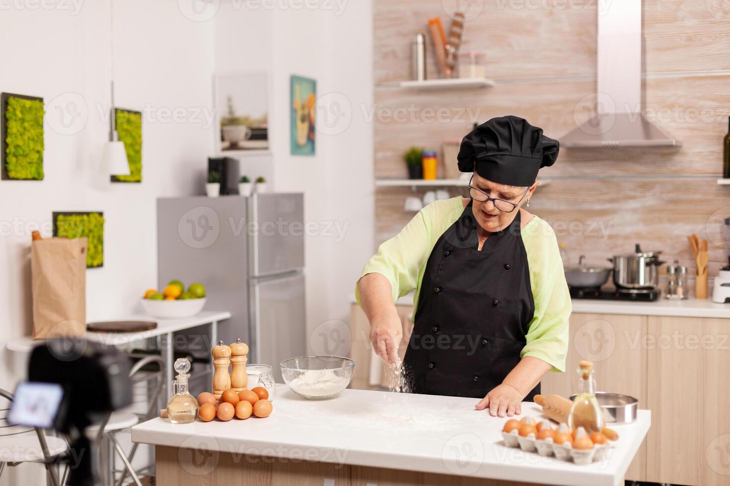 Senior woman creating content for culinary blog preparing tasty bread. Retired blogger chef influencer using internet technology communicating, shooting blogging on social media with digital equipment. photo