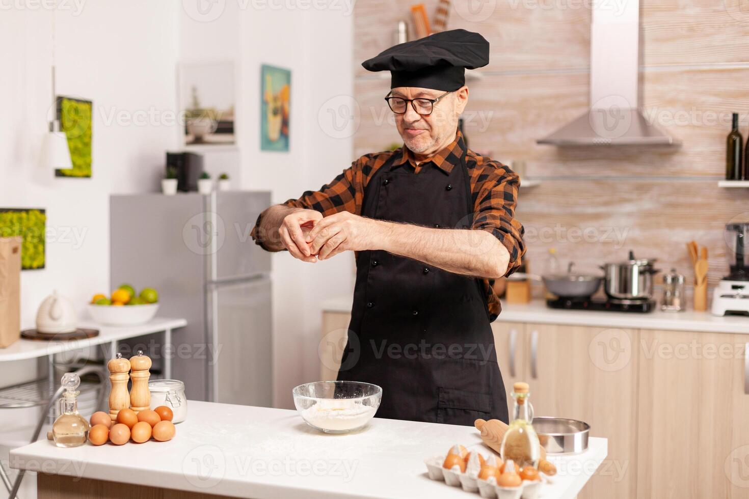 Elderly man cracking eggs over wheat flour while preparing delicious food. Elderly pastry chef cracking egg on glass bowl for cake recipe in kitchen, mixing by hand, kneading ingredients prreparing homemade cake photo