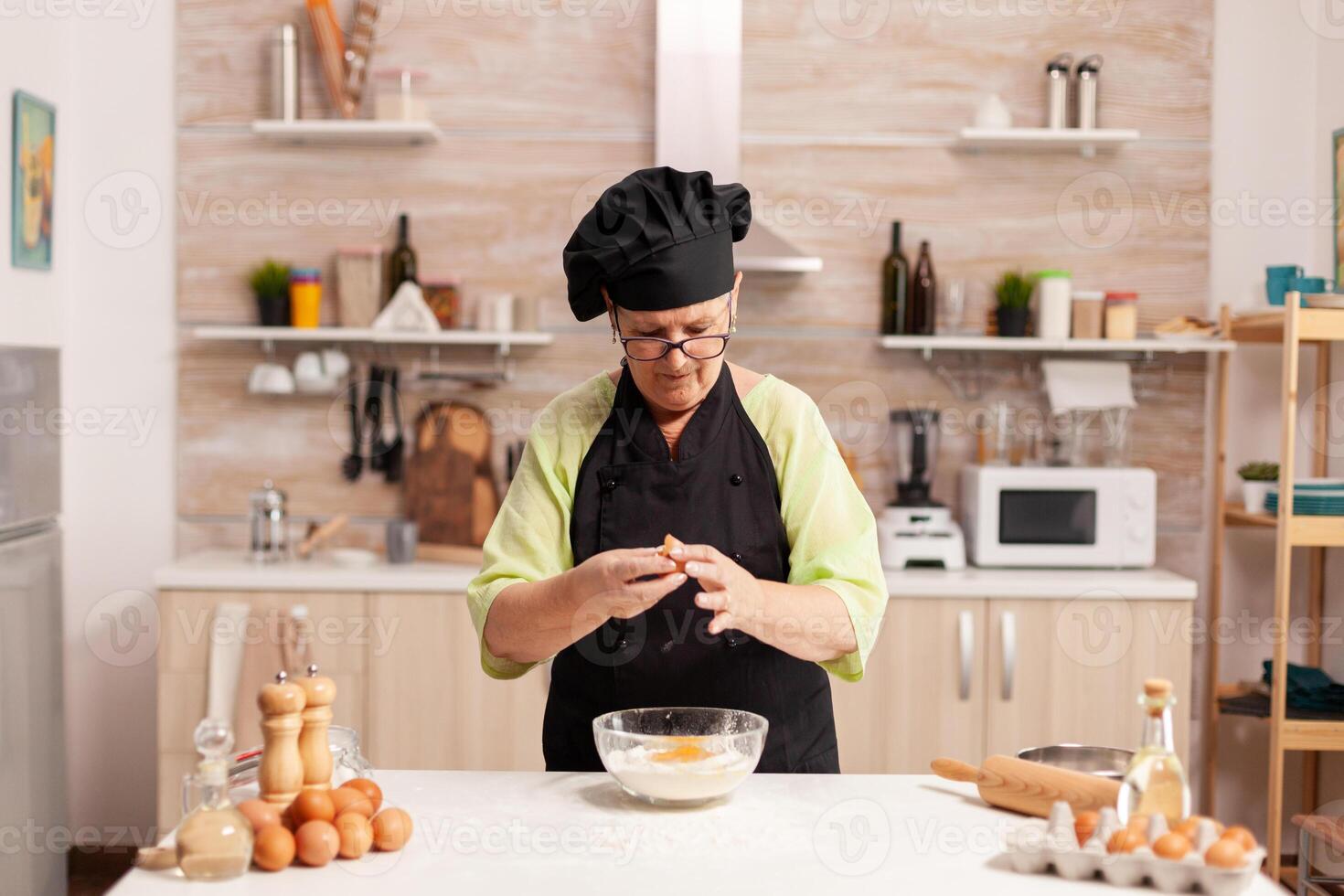 Chef hands with flour and eggs in preparation process for baking. Elderly pastry chef cracking egg on glass bowl for cake recipe in kitchen, mixing by hand, kneading. photo