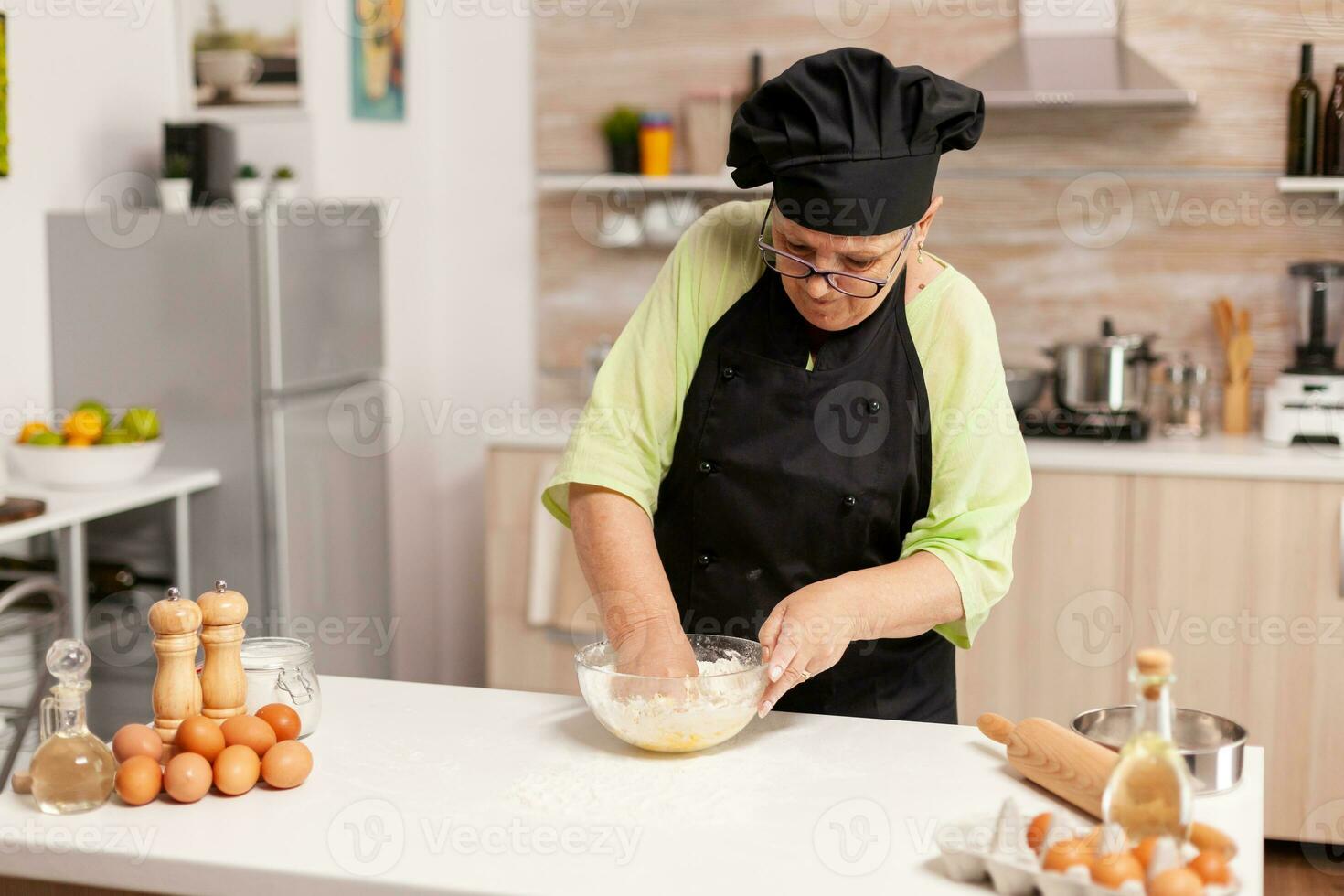 Chef in home kitchen preparing pasta dough following traditional recipe. Retired elderly chef with uniform sprinkling, sieving sifting raw ingredients and mixing . photo