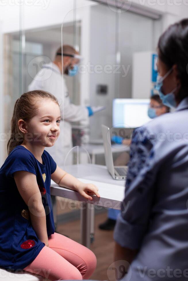 Mother talking with daughter about treatment in hospital clinic. Physician specialist in medicine with face mask and visor against covid19 providing professional services photo