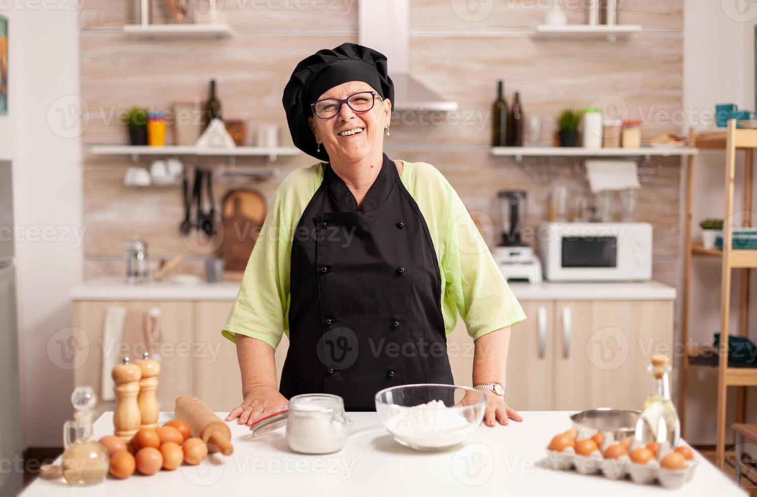 Happy elderly woman wearing bonete looking at camera in dining room at home. Retired elderly baker in kitchen uniform preparing pastry ingredients on wooden table ready to cook homemade tasty bread. photo