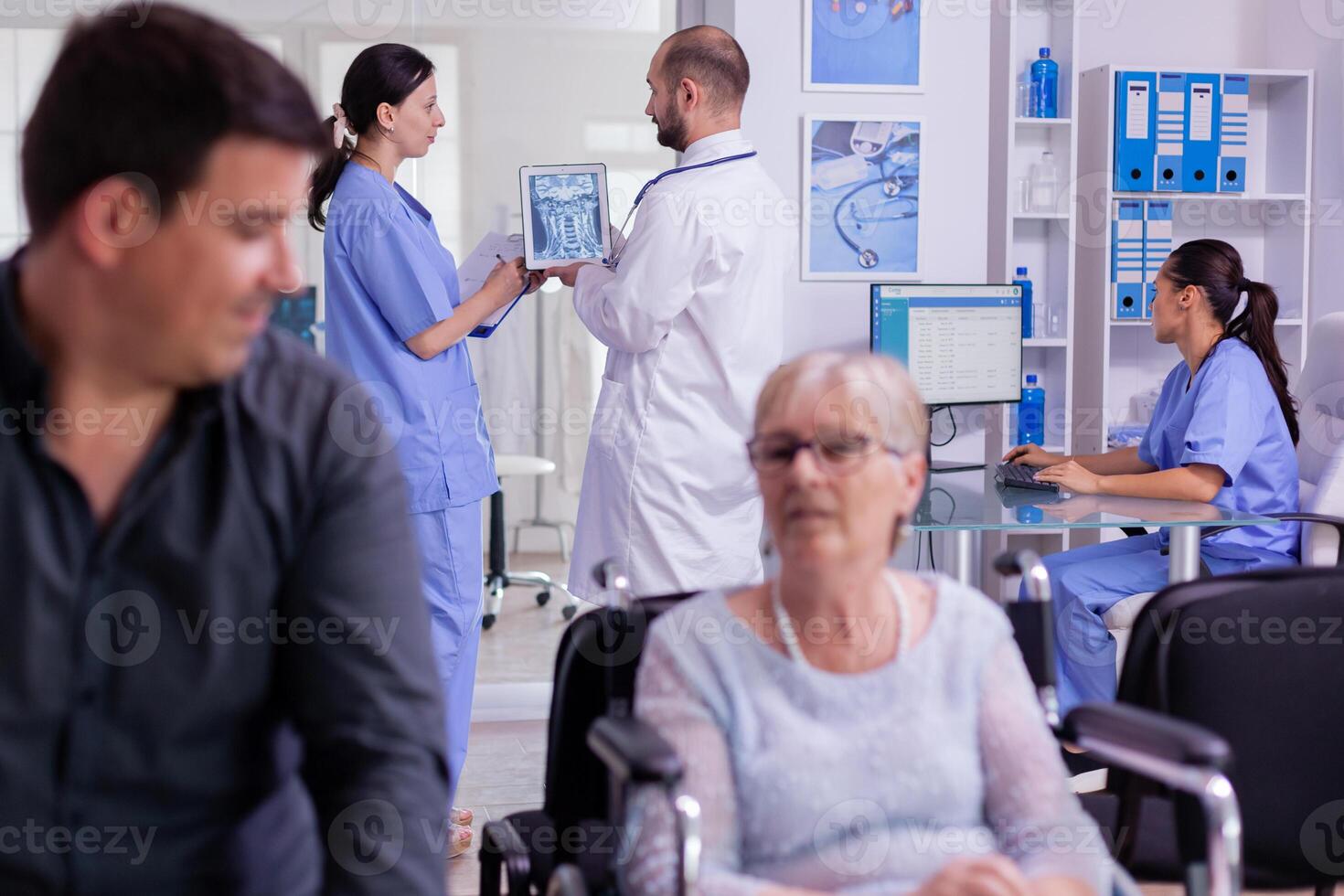 Doctor holding patient x-ray explaining diagnosis to nurse in waiting area. Disabled senior woman wheelchair waiting for medical examination. Hospital and health care system, medicine private clinic photo