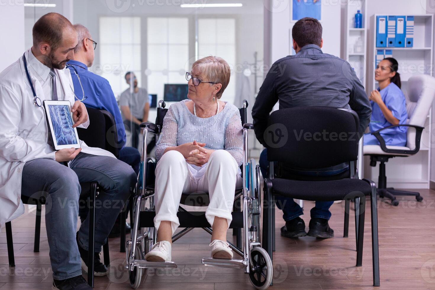 Doctor holding tablet pc with x-ray while explaining diagnosis to disabled elderly woman in wheelchair. Handicapped patient in hospital waiting area. Man in examination room. photo