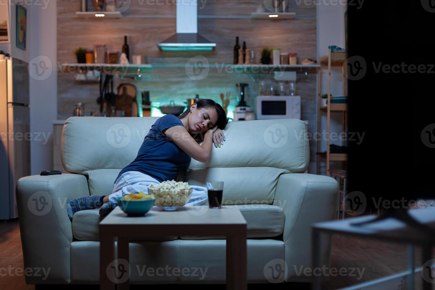 Young tired after work woman looking asleep in the evening in front of TV. Exhausted lonely sleepy lady in pajamas sleeping on sofa while watching a bored movie in living room, closing eyes at night photo