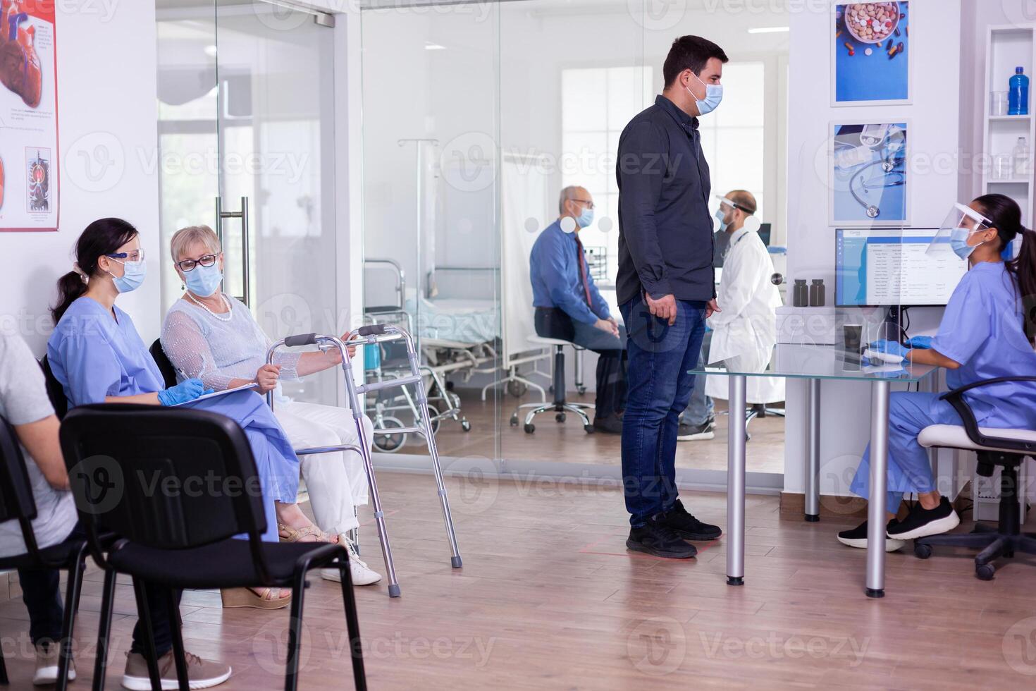 Young man wearing face mask against coronavirus while waiting at hospital reception, talking with nurse. Invalid mature woman with walking frame talking with doctor assistant before consultation photo