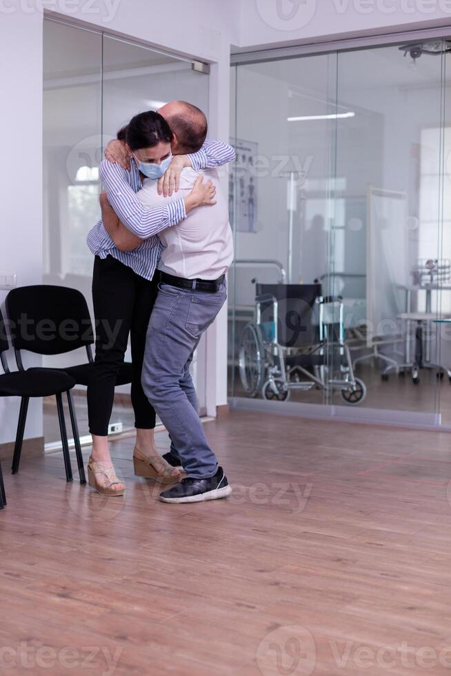 Happy young couple after receiving good from doctor wearing protection face mask in hospital waiting area hugging eatch other. Positive man and woman in clinic after test results during global pandemic photo