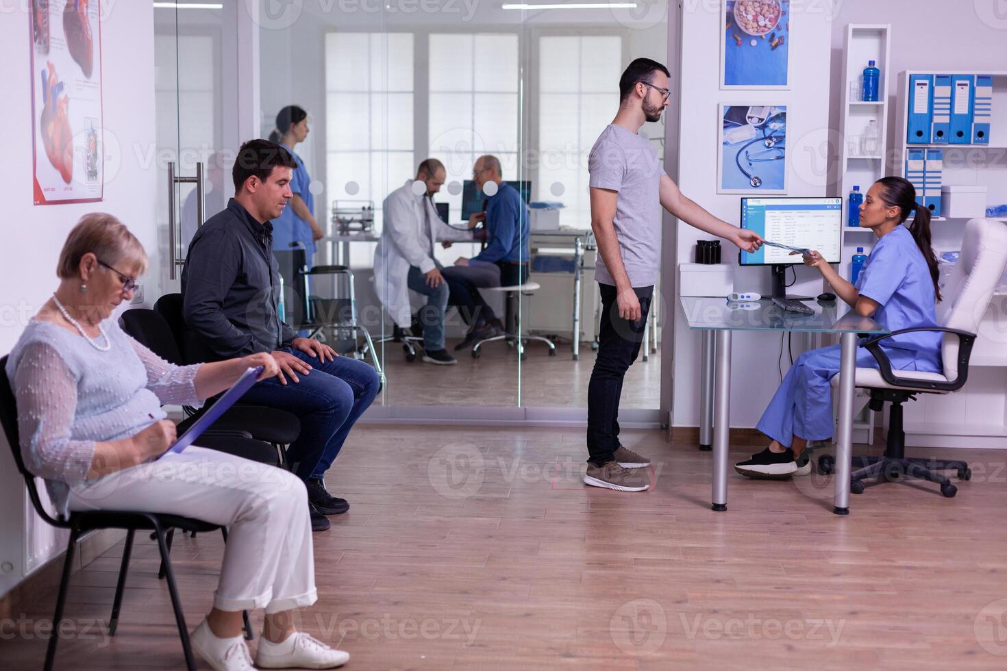 Man visiting hospital clinic for medical checking, giving radiography to assistant for new appointment at specialist doctor. Patients sitting in crowded waiting room filling form for consultation photo