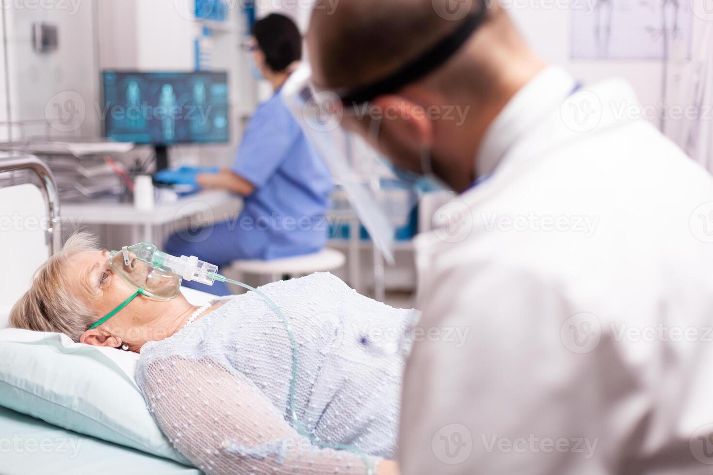 Doctor checking senior woman with oxygen mask breathing hard because of infection with covid19. Patient in hospital room laying on medical bed with infected lung during coronavirus pandemic having respiratory problem. photo