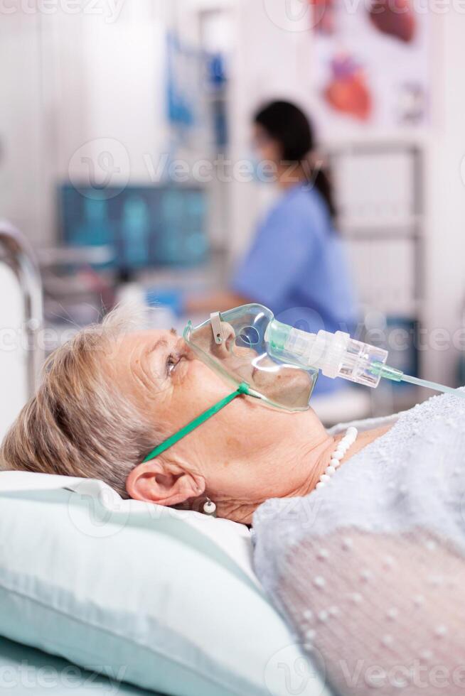 Senior woman contaminated with covid19 wearing oxygen mask in clinic. Patient in hospital room laying on medical bed with infected lung during coronavirus pandemic having respiratory problem. photo