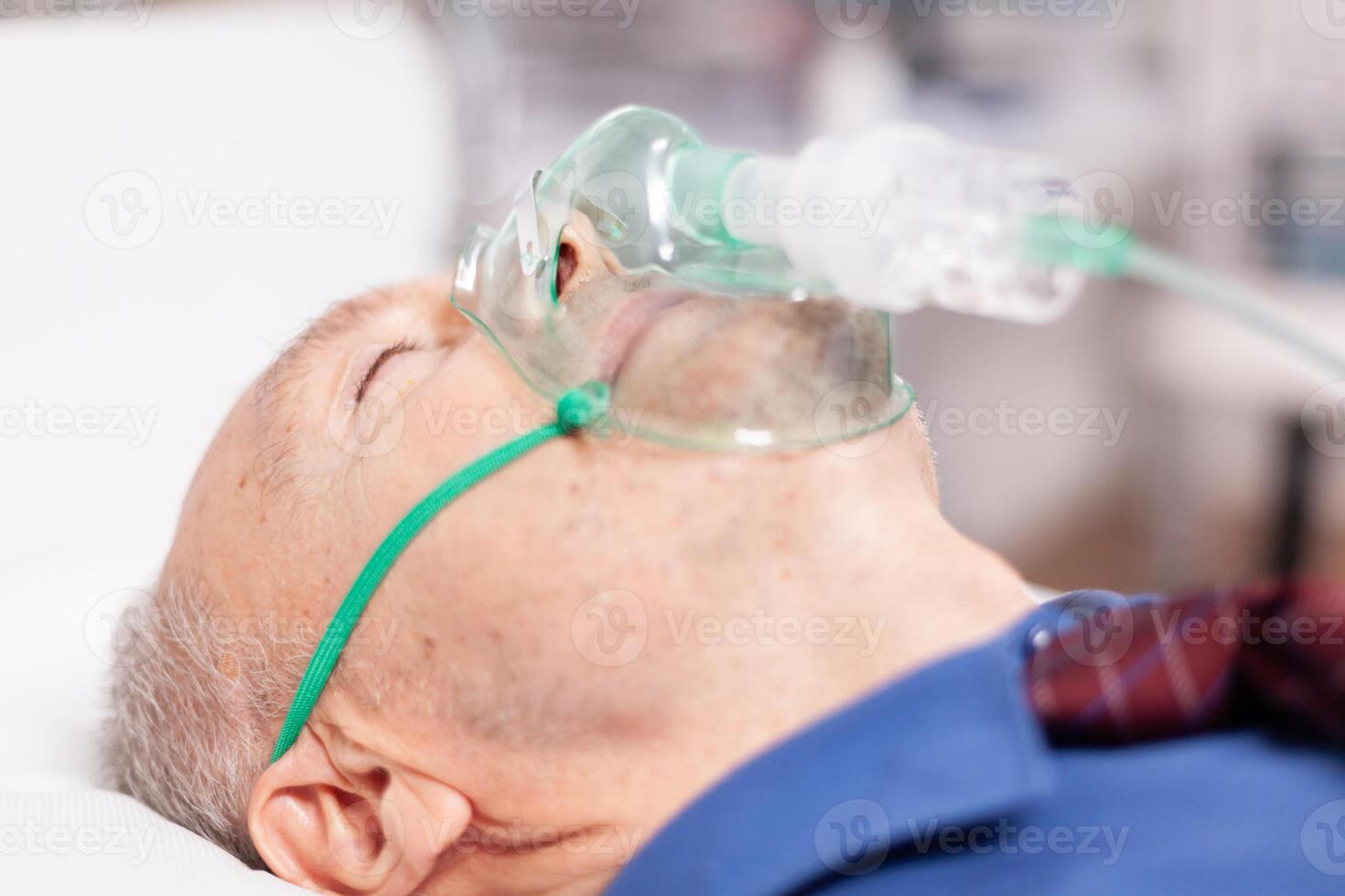 Senior man doing inhalation through oxygen mask because of covid19. Patient in hospital room laying on medical bed with infected lung during coronavirus pandemic having respiratory problem. photo