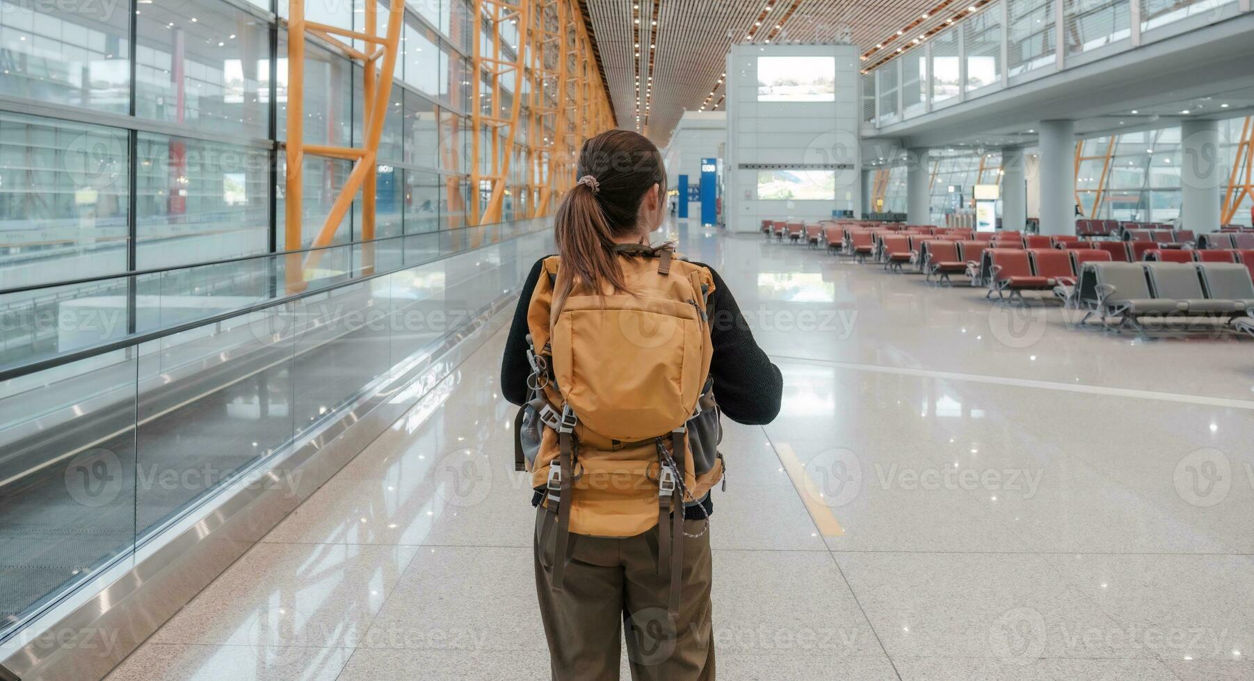 A young Asian woman stands behind in casual clothes and carries a backpack. The schedule can be found on the airport station's scoreboard at weekend. photo