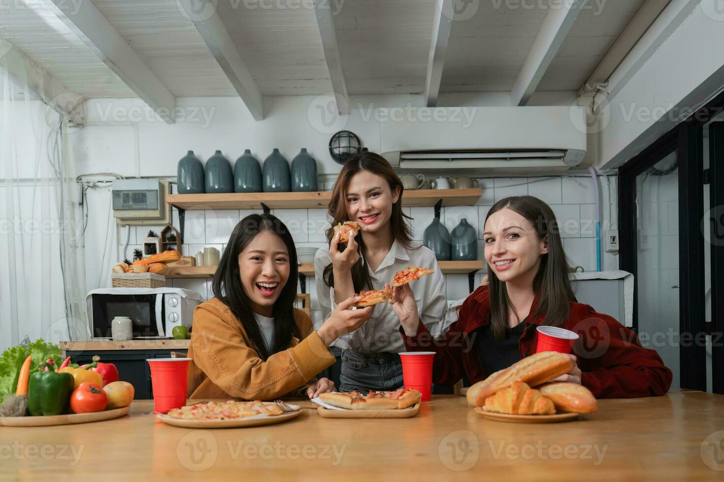 A group of young female friends have a party with pizza on the table and red drink glasses. Talk and live together happy, having fun at home. photo
