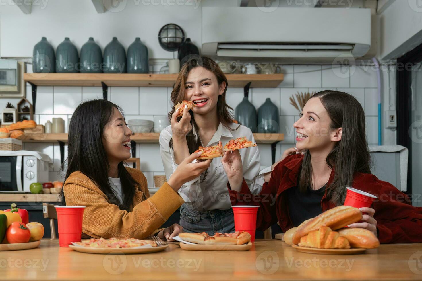 A group of young female friends have a party with pizza on the table and red drink glasses. Talk and live together happy, having fun at home. photo