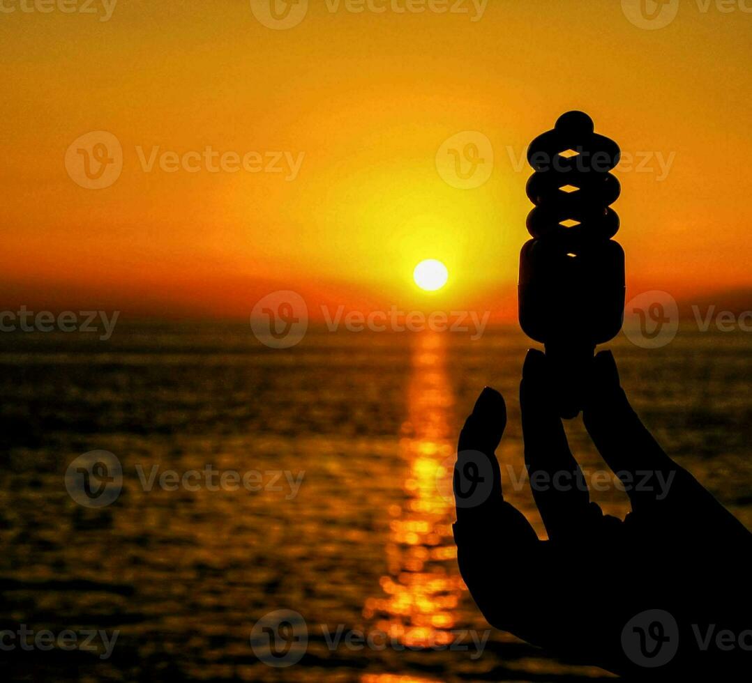 a person holding up a light bulb in front of the sunset photo