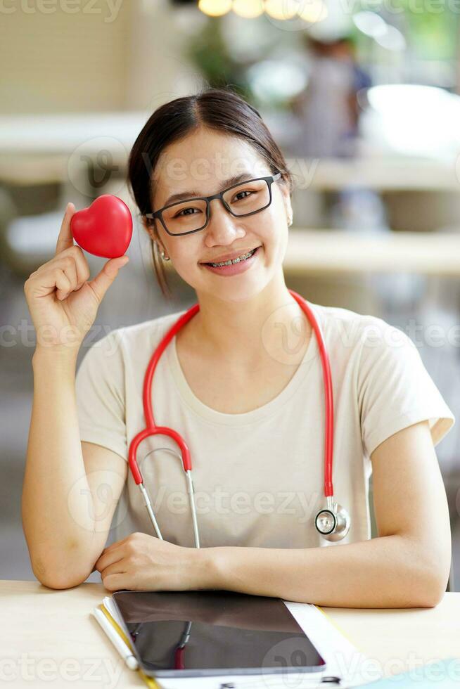 Medical student action holding a rubber red heart with cute smile on blurred background. Asian medical student with Heart disease concept. photo
