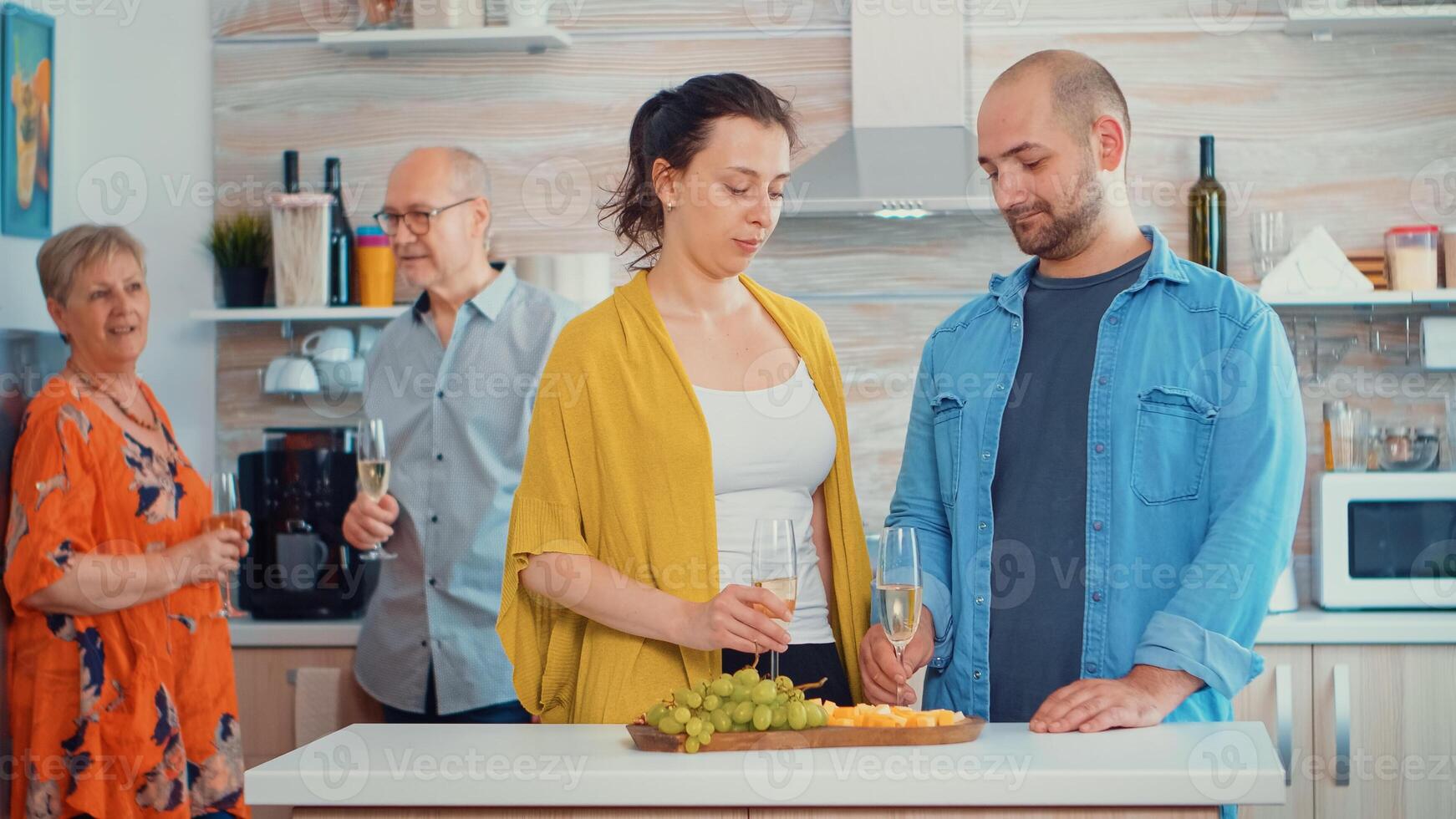 Handsome man making proposal to his girlfriend in front of his parents, in kitchen sitting near the table drinking a glass of white wine. Happy surprised woman kissing him while parents take a photo