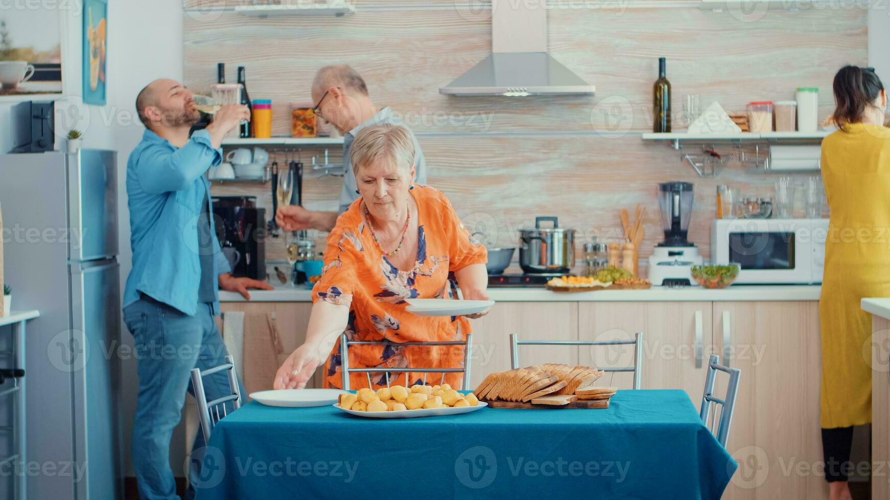 Middle aged woman and older senior have fun working together setting the dinner table in kitchen, while men talking in background and drinking a glass of white wine during a relaxing family day. photo