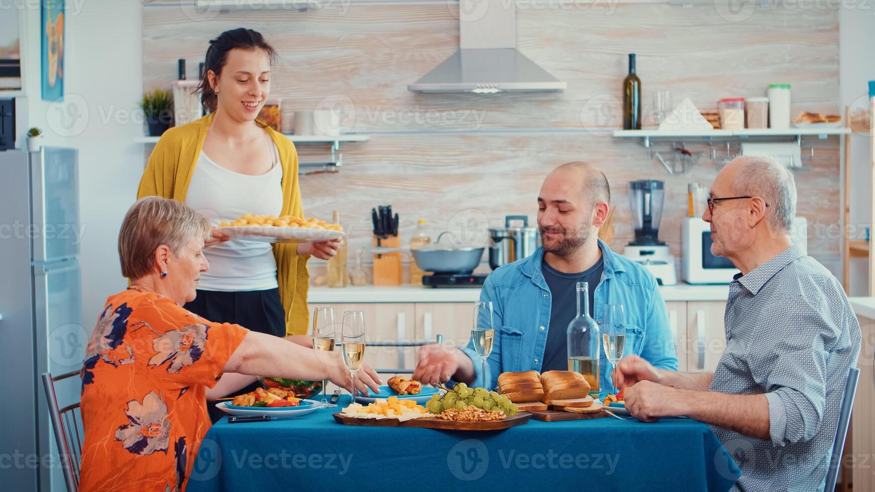Young wife putting potatoes on the table and clinking a glass of wine with her husband. Caucasian family enjoying time at home, in the kitchen sitting by the table, eating dinner together and drinking photo
