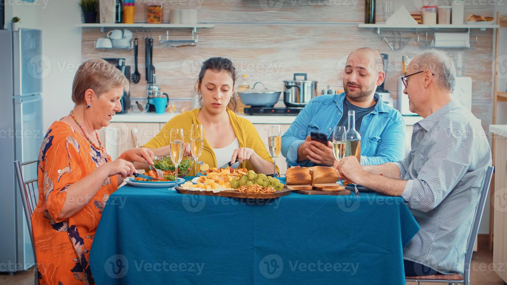 Man using phone during dinner showing some pictures to her mother. Multi generation, four people, two happy couples talking and eating during a gourmet meal, enjoying time at home. photo