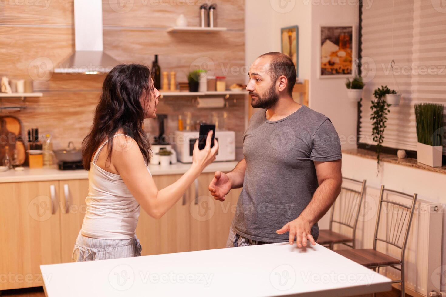 Woman looking disappointed at husband after catching him cheating while holding phone. Heated angry frustrated offended irritated accusing her man of infidelity showing him messages . photo