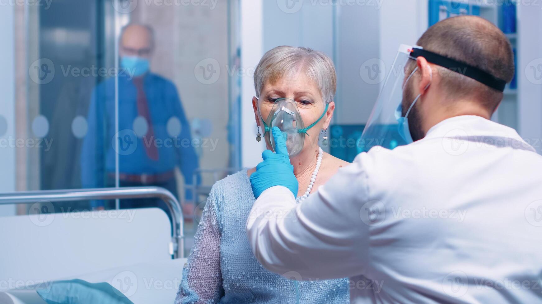 Doctor in mask and visor helping old lady to breathe with a respiratory oxygen mask while sitting on hospital bed in modern private clinic. Coronavirus covid-19 outbreak healthcare crisis. photo