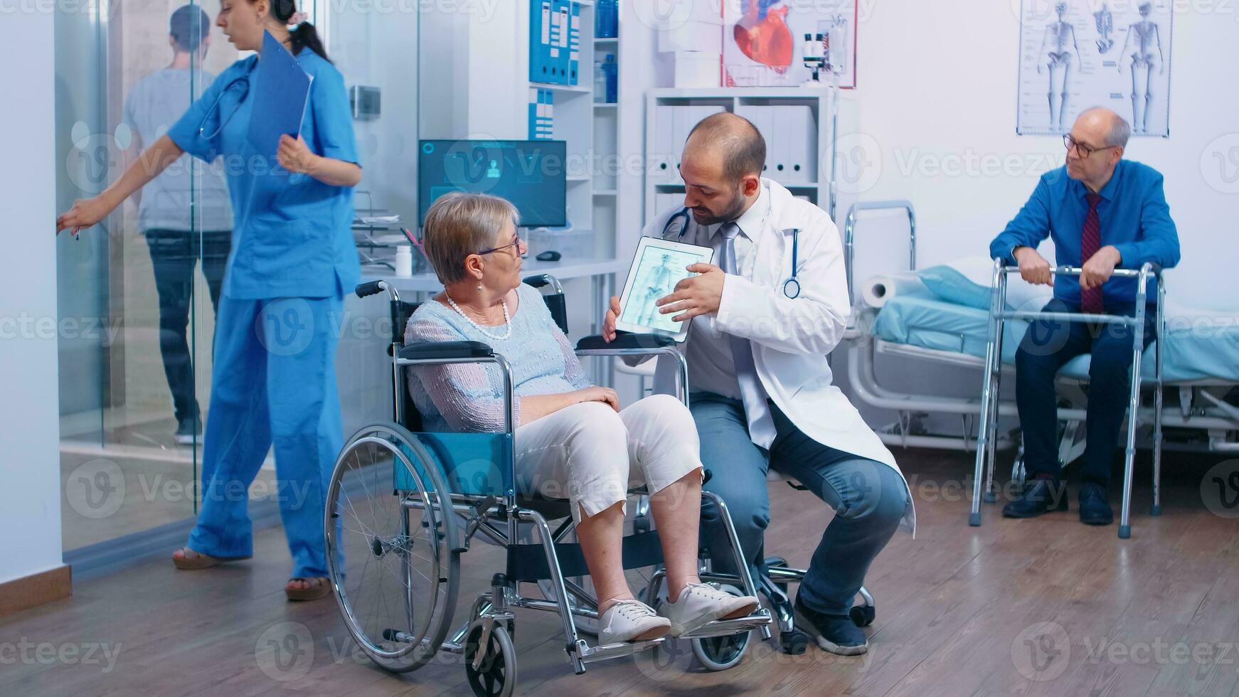 Doctor showing the osteoporosis risk for elderly persons to a senior woman in wheelchair in a rehabilitation clinic, center or hospital. Old man with walking frame, health care medical facility, modern medicine photo