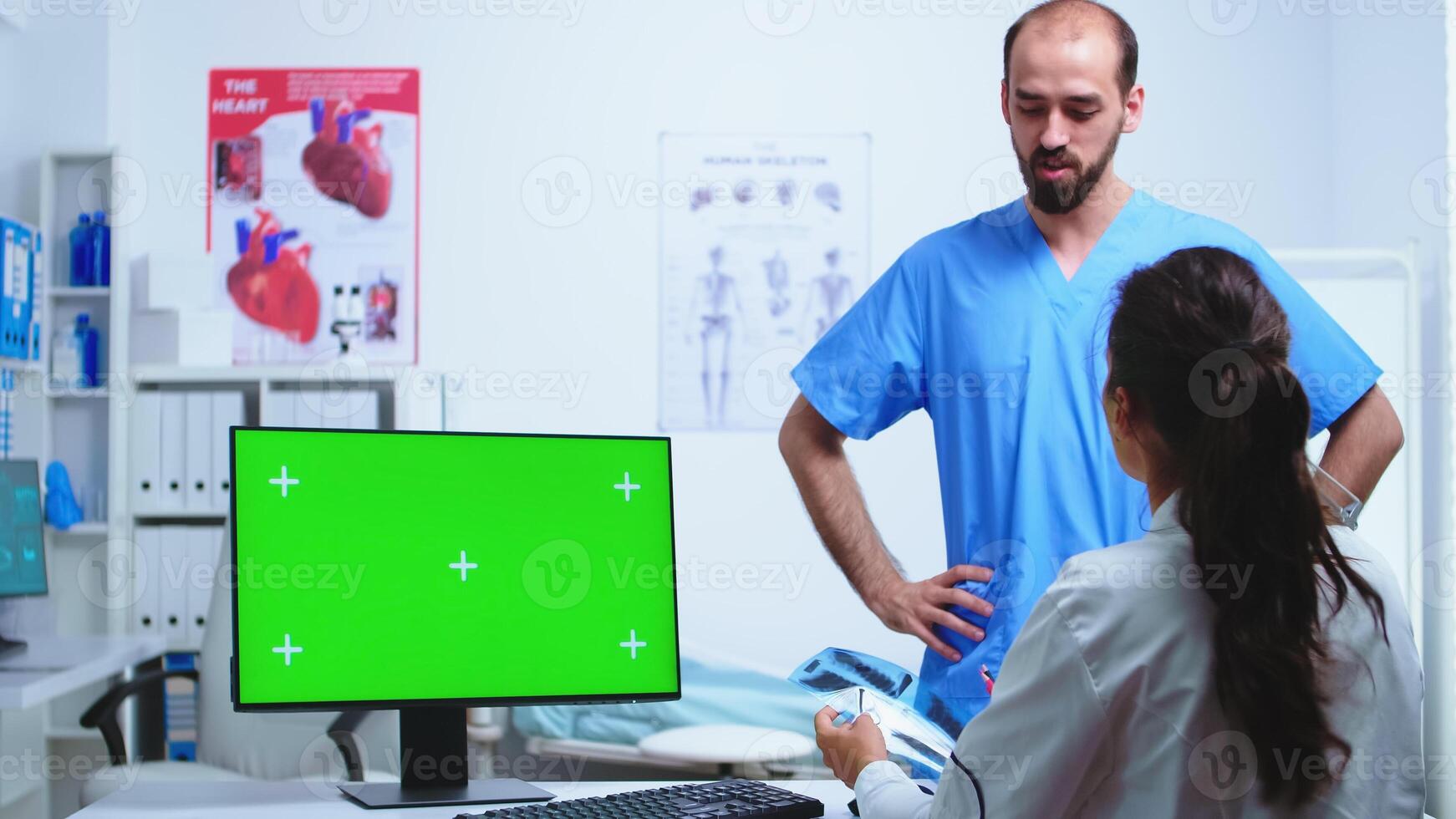 Assistant giving doctor x-ray image while working on computer with green screen monitor in hospital cabinet. Desktop with replaceable screen in medical clinic while doctor is checking patient radiography for diagnose. photo