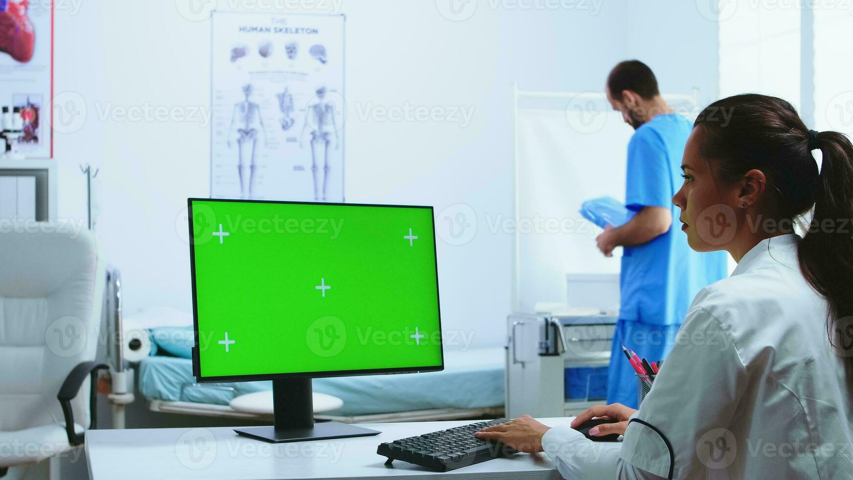 médico utilizando computadora con verde pantalla monitor en hospital gabinete y asistente en el antecedentes. médico en blanco Saco trabajando en monitor con croma llave en clínica gabinete a cheque paciente diagnosticar. foto