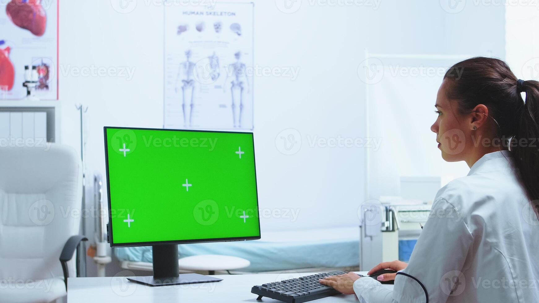 Chroma key green screen on computer in hospital cabinet while medic is working on it. Nurse in blue uniform in the background. Computer with repleceable screen used by medicine specialist in hospital and wearing uniform. photo